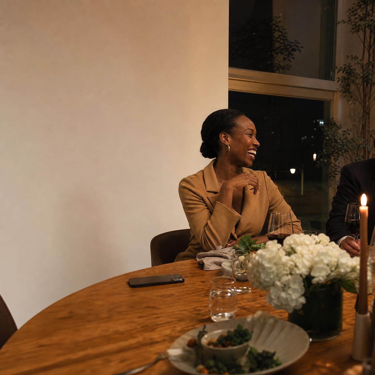 A Black woman in a camel blazer at a warm dinner table, mid-laugh, her phone lying face-down and forgotten near the edge of the table.