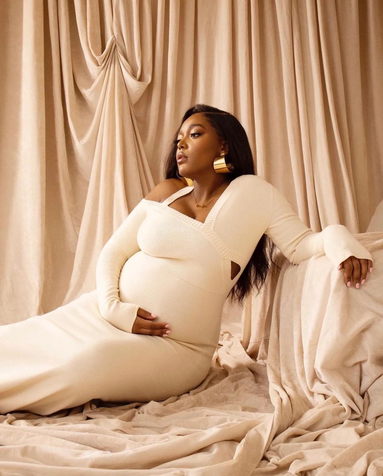 Anifa Mvuemba, pregnant, seated in a cream gown against cream drapery with one hand resting at her jaw.