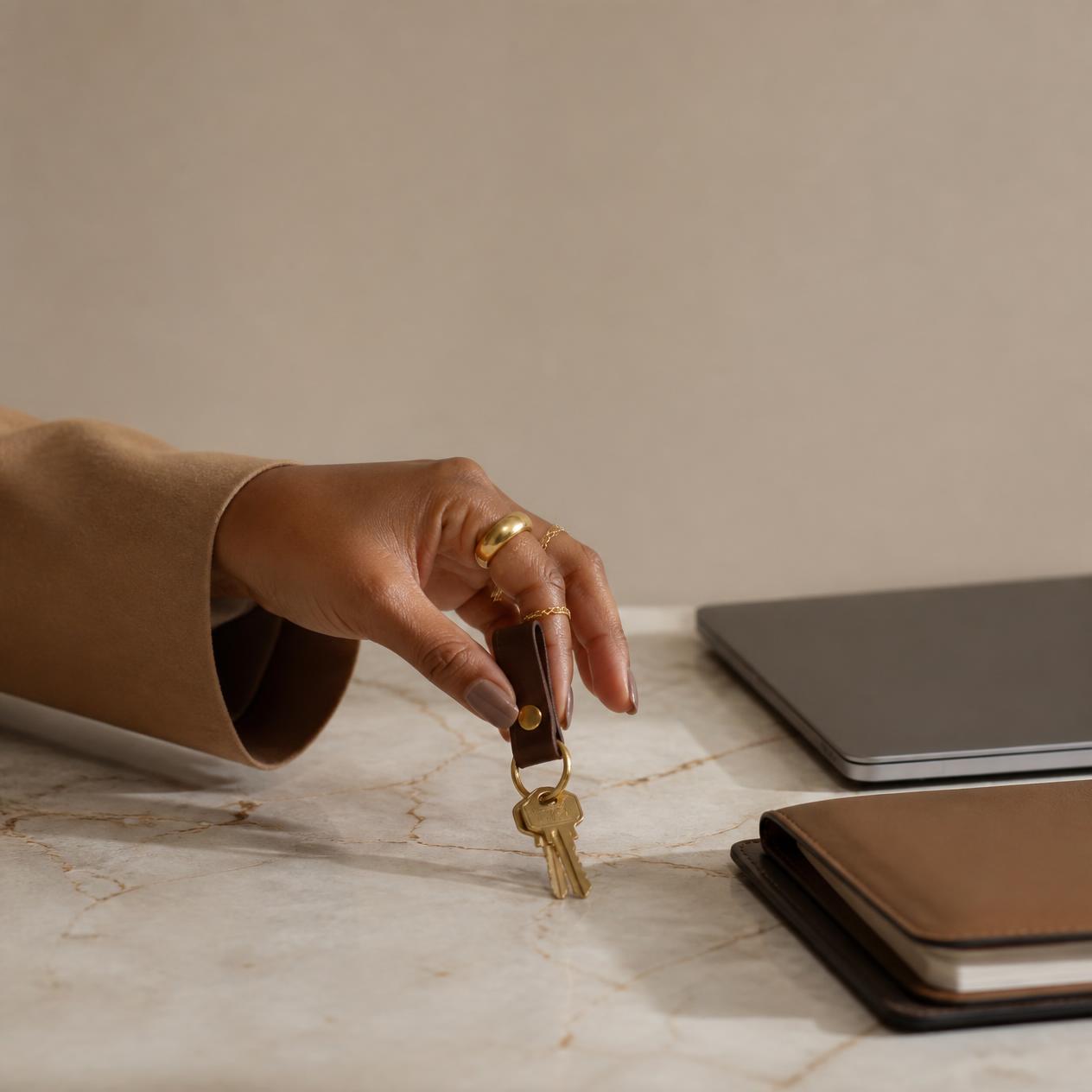 A Black woman's hand, manicured with stacked gold rings, lifting a single set of keys off a marble counter beside an unbranded laptop and a slim folder.