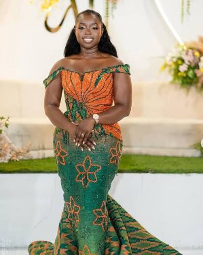 Ama Hill in an embroidered orange and green gown, hands clasped in front of her, smiling at a formal event.