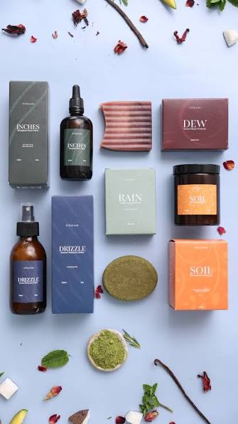 A flat-lay of Plantmade hair and skincare products β Inches, Dew, Rain, Soil, and Drizzle β arranged with dried herbs, vanilla pods, and rose petals.