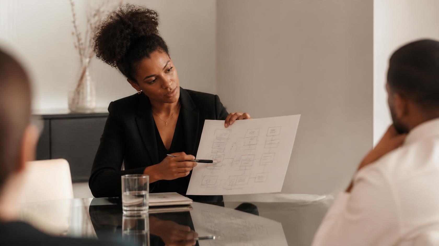 A Black woman in a black blazer at a client's glass conference table, confidently pointing to a clean, organized system diagram.
