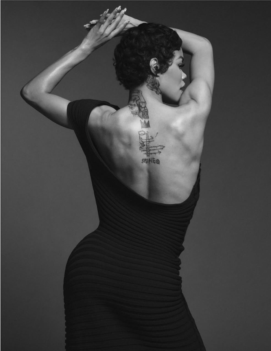 A black-and-white portrait of Teyana Taylor from behind, arms raised, in a fitted black gown, a small tattoo visible on her upper back.