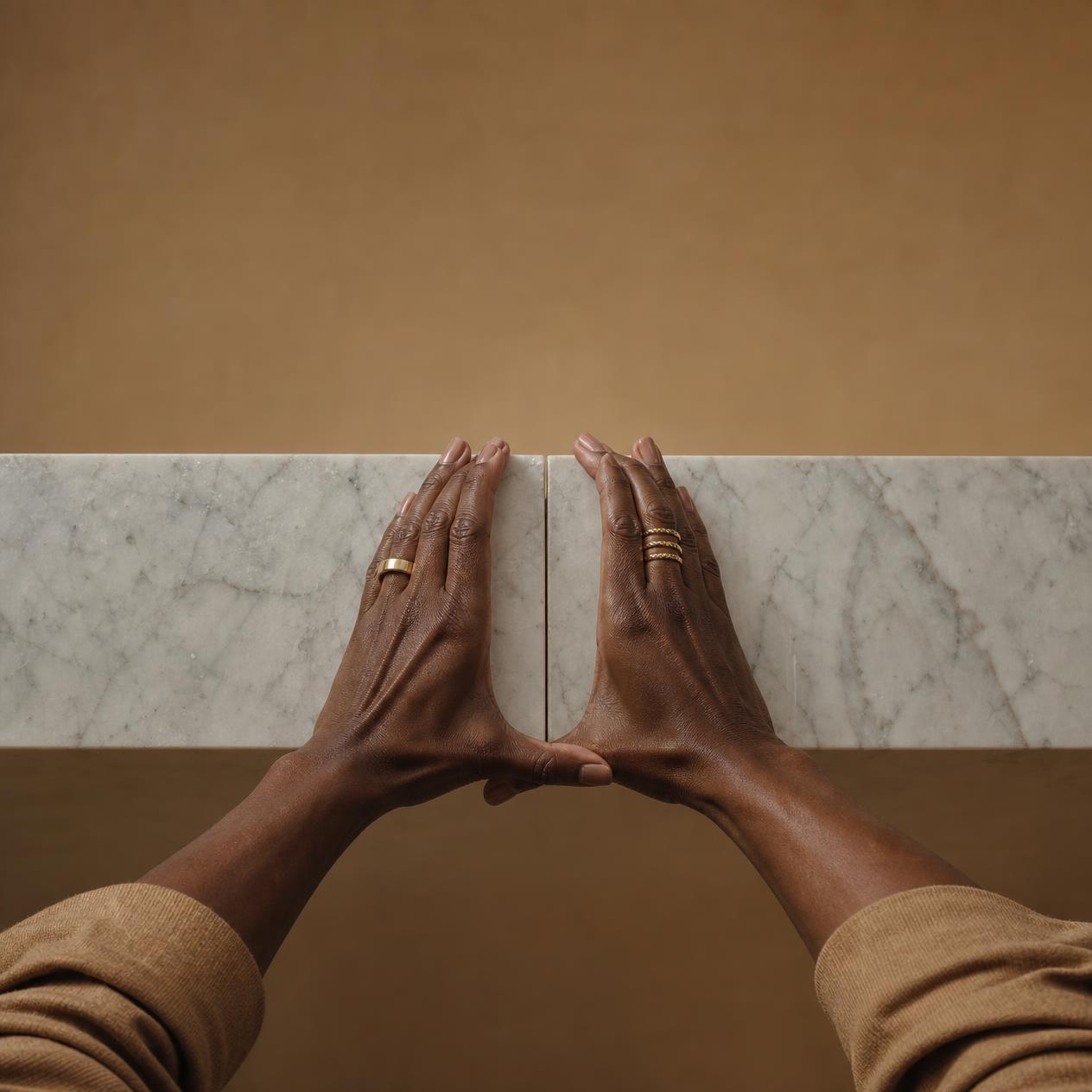 A Black woman's hands, manicured with stacked gold rings, physically holding two heavy marble slabs together at the seam, arms braced under the full weight.