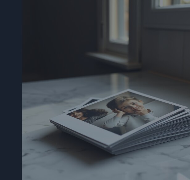 A stack of printed family photographs on a marble counter, slightly fanned, daylight falling across them, one edge visibly curling.