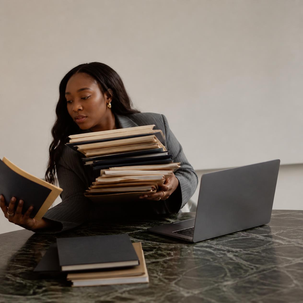 A Black woman in a grey suit balancing an overloaded stack of folders beside an open laptop on a marble desk.